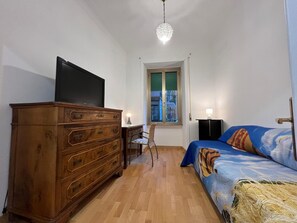 A bedroom with a wooden dresser, a television, a bed with a colorful headboard, a window with a view, and a chandelier.