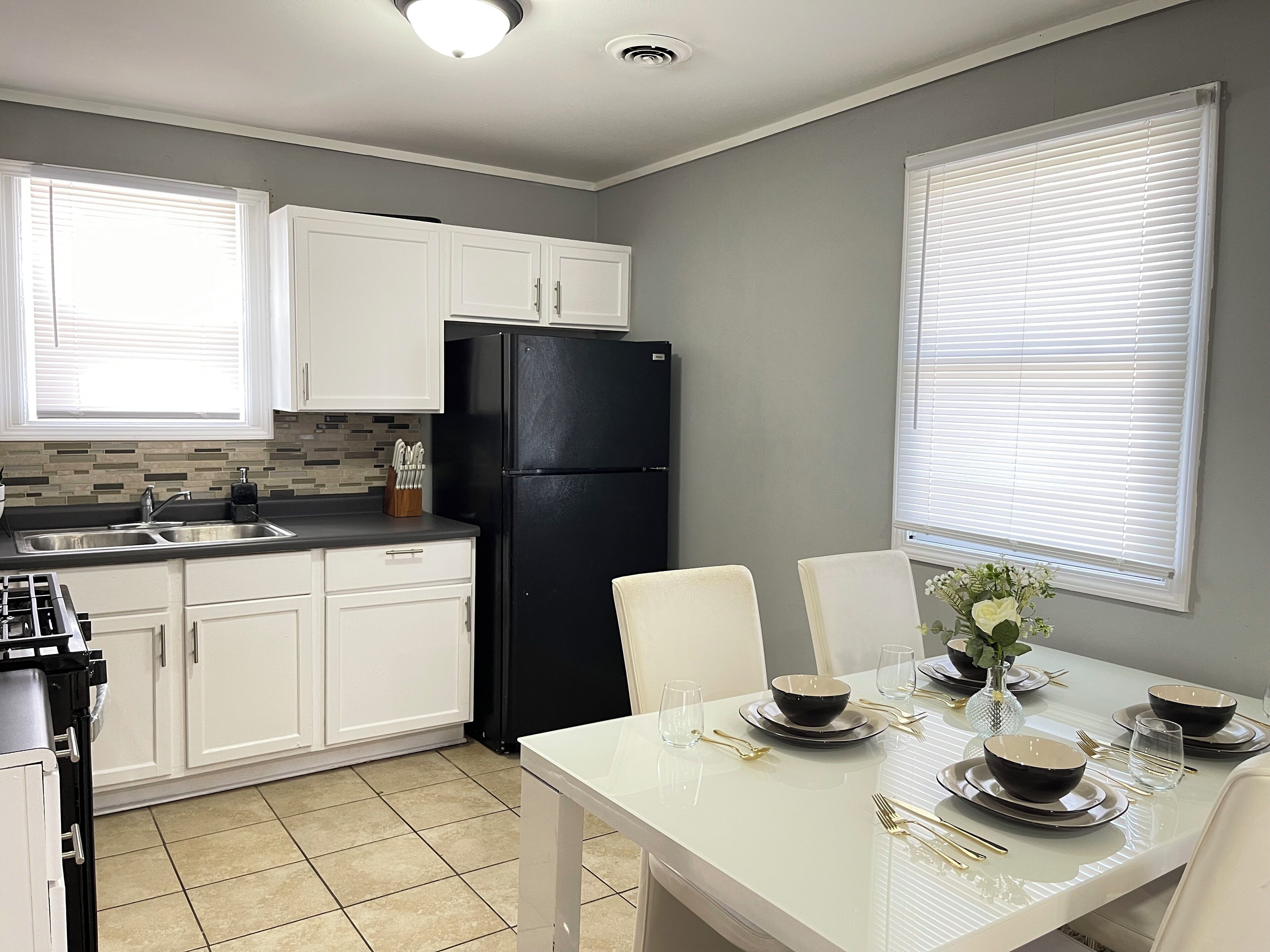 Une cuisine moderne avec des meubles blancs, un réfrigérateur noir, un pare-feu en carrelage et un coin repas aménagé pour quatre personnes, avec une table et des chaises blanches.