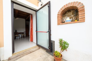 Une porte vitrée ouverte donne sur une pièce où se trouvent une table et des chaises, une plante en pot et un four en briques encastré dans le mur.