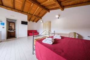 Quarto com teto de madeira, cama com colcha vermelha, parede branca e porta que dá para um banheiro.