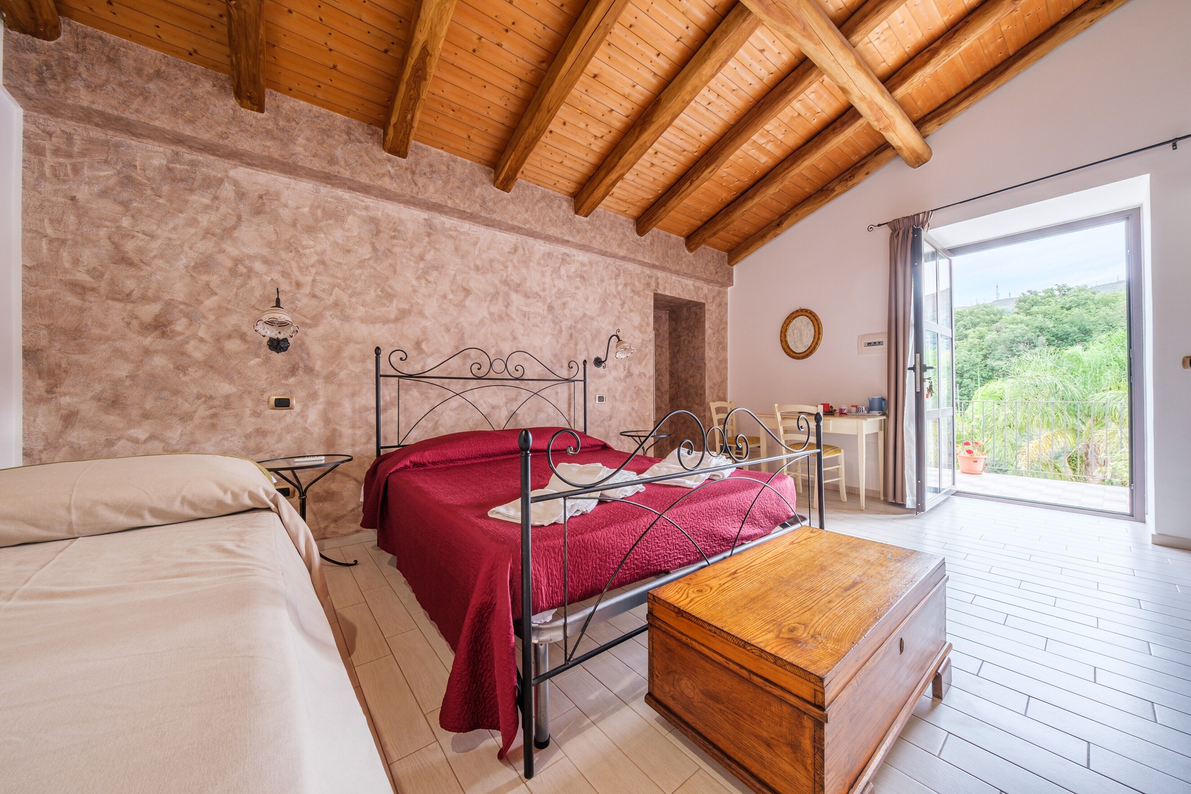 Un dormitorio con cama, un cofre de madera, un escritorio y vista a la vegetación a través de un amplio ventanal.