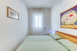 A bedroom with a bed, a nightstand, a lamp, a window with curtains, and a wall painting. - Apartment 'Trande' with Balcony, Wi-Fi and Air Conditioning (Santa Maria di Leuca)