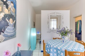 Een eetruimte met een tafel gedekt voor twee personen, een vaas met bloemen, een schilderij aan de muur en een hoge blauwe lamp.
