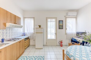Een kleine keuken met een kookplaat, gootsteen en koelkast, een eethoek met een tafel en stoelen, en een deur naar een andere kamer.