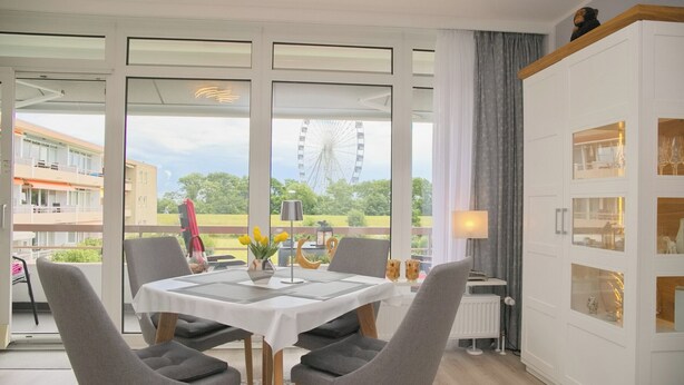A dining area with a table and chairs, large windows, and a view of a Ferris wheel. - House Baltic App. 97 incl. WLAN (Grömitz)