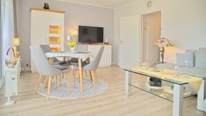A modern living room with a glass coffee table, a dining area with a white table and grey chairs, a flat-screen TV mounted on a wooden cabinet, and a sofa with a floral lamp. - House Baltic App. 97 incl. WLAN (Grömitz)