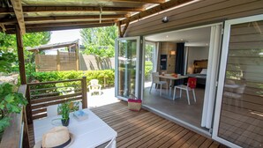 A wooden deck with a table and chairs, a sliding glass door leading to a dining area, and a covered seating area with a hat and plant on the table. - Camping international Aups 83 (Aups)