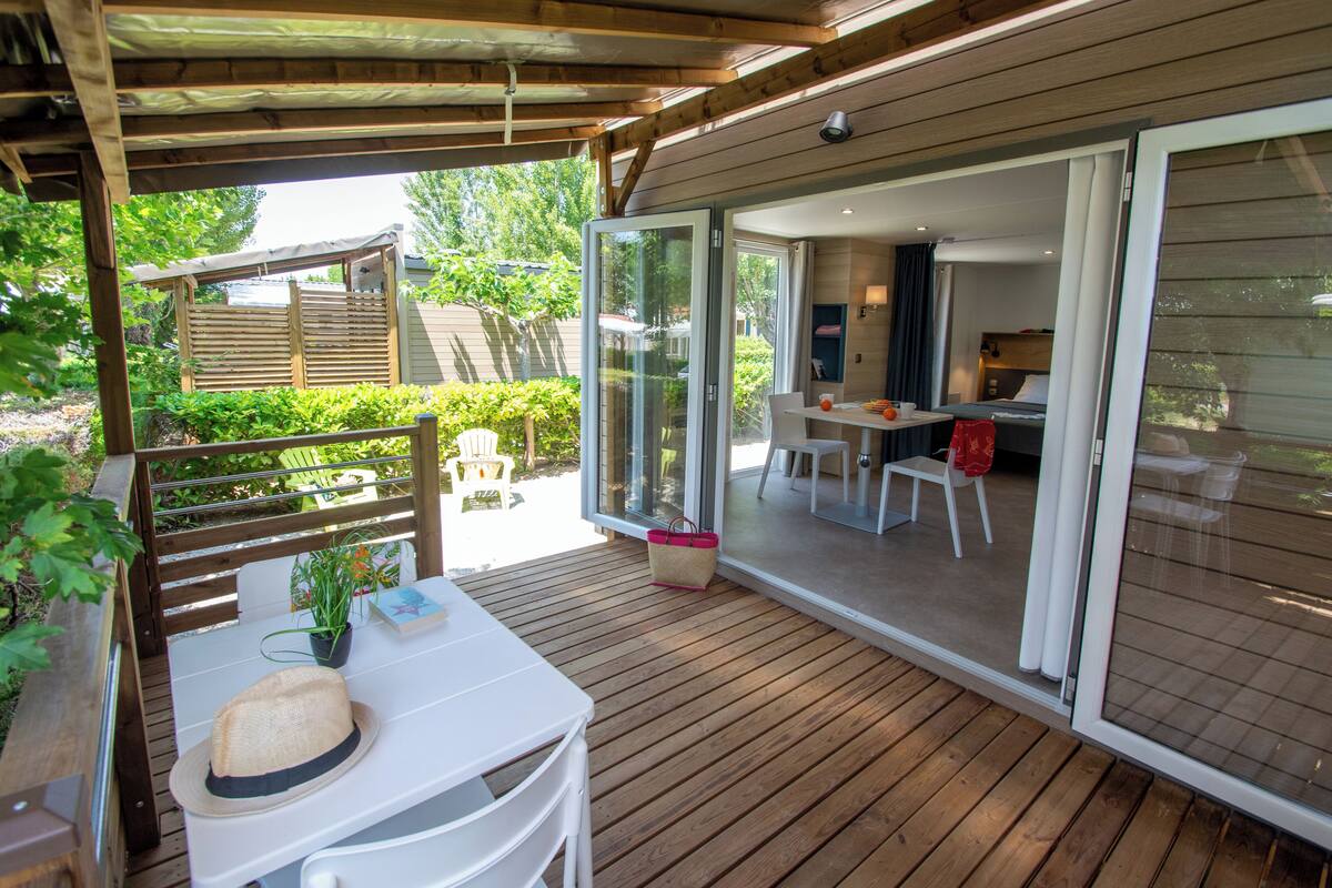 A wooden deck with a table and chairs, a sliding glass door leading to a dining area, and a covered seating area with a hat and plant on the table.