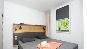 A modern bedroom with a wooden headboard, two bedside lamps, a window with a view of greenery, and a neatly made bed with a striped blanket and a hat. - Camping international Aups 83 (Aups)