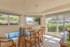 Un espace repas avec une table en verre et des chaises en bois, de grandes fenêtres donnant sur la verdure et les bâtiments, et un luminaire au plafond.