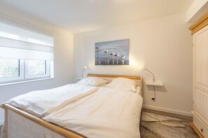 Een slaapkamer met een houten bed, wit beddengoed, een schilderij van vogels aan de muur, een nachtkastje met een lamp en een raam met jaloezieën.