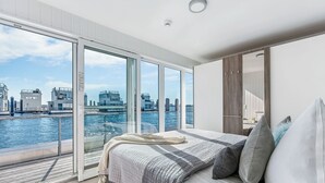 A modern bedroom with a large bed, a view of the city through floor-to-ceiling windows, and a balcony overlooking a body of water.