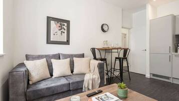 En stue med en grå sofa, et sofabord med en plante, et indrammet billede på væggen, og et spiseområde med bord og stole.