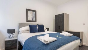 A neatly made bed with a blue quilt, two pillows, and a folded towel on a white mattress. There is a nightstand with a lamp and a drawer, and a wardrobe with a dark door.