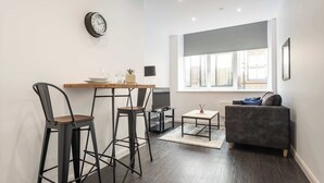 A modern living room with a grey sofa, a wooden coffee table, and a TV on a stand. There are two black bar stools and a small table with a clock and decorative items.
