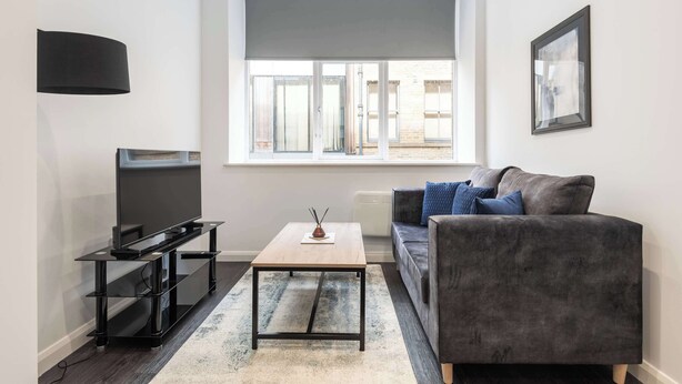 A modern living room with a flat-screen TV on a black stand, a wooden coffee table, a grey sofa with blue cushions, and a large window with a grey roller blind.