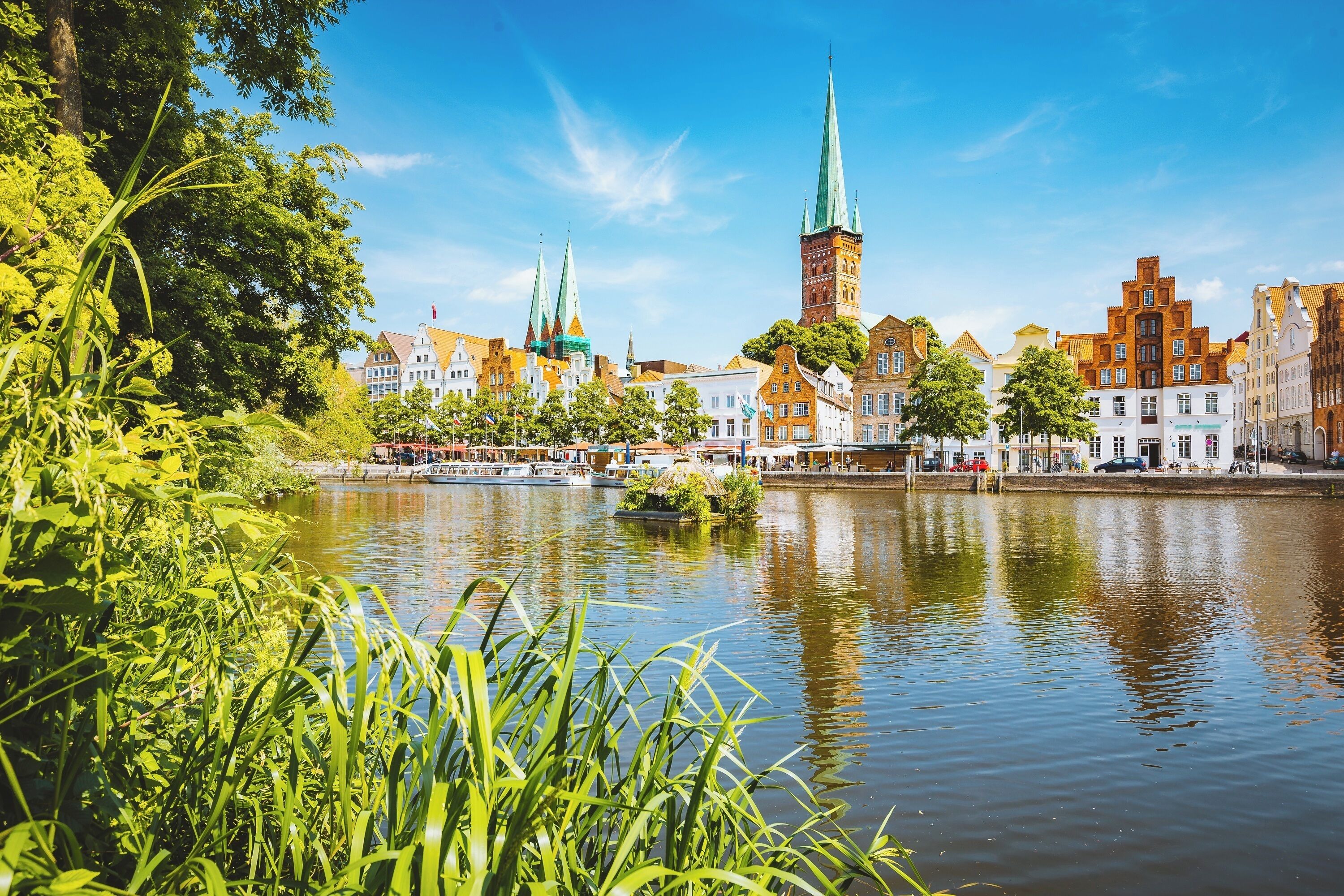 Een uitzicht op de rivier met historische gebouwen, een opvallende toren en spitsen, omringd door weelderig groen.