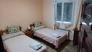 Una habitación con dos camas, un ventilador de techo, una ventana con persianas, un radiador y un aparador de madera.