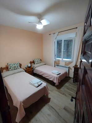 Una habitación con dos camas, un ventilador de techo, una ventana con persianas, un radiador y un aparador de madera.