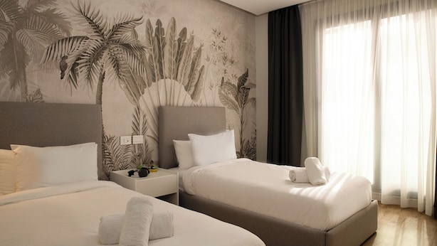 Quarto de hotel com duas camas, uma mesinha de cabeceira, uma luminária e uma janela grande com cortinas.