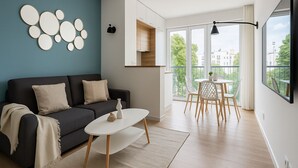 A modern living room with a dark sofa, a white coffee table, and a dining area with a table and chairs. There is a large window with a view of buildings and greenery. - Charmant T2 lumineux près de Montmartre (Paris)