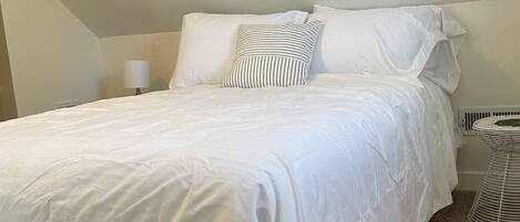 A neatly made bed with white bedding and striped pillows, a wooden headboard, a bedside lamp, and a radiator in a room with beige walls and carpet.