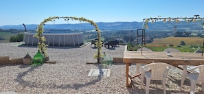 Um terraço no telhado com mesa e cadeiras de madeira, um arco decorativo e vista para o campo.