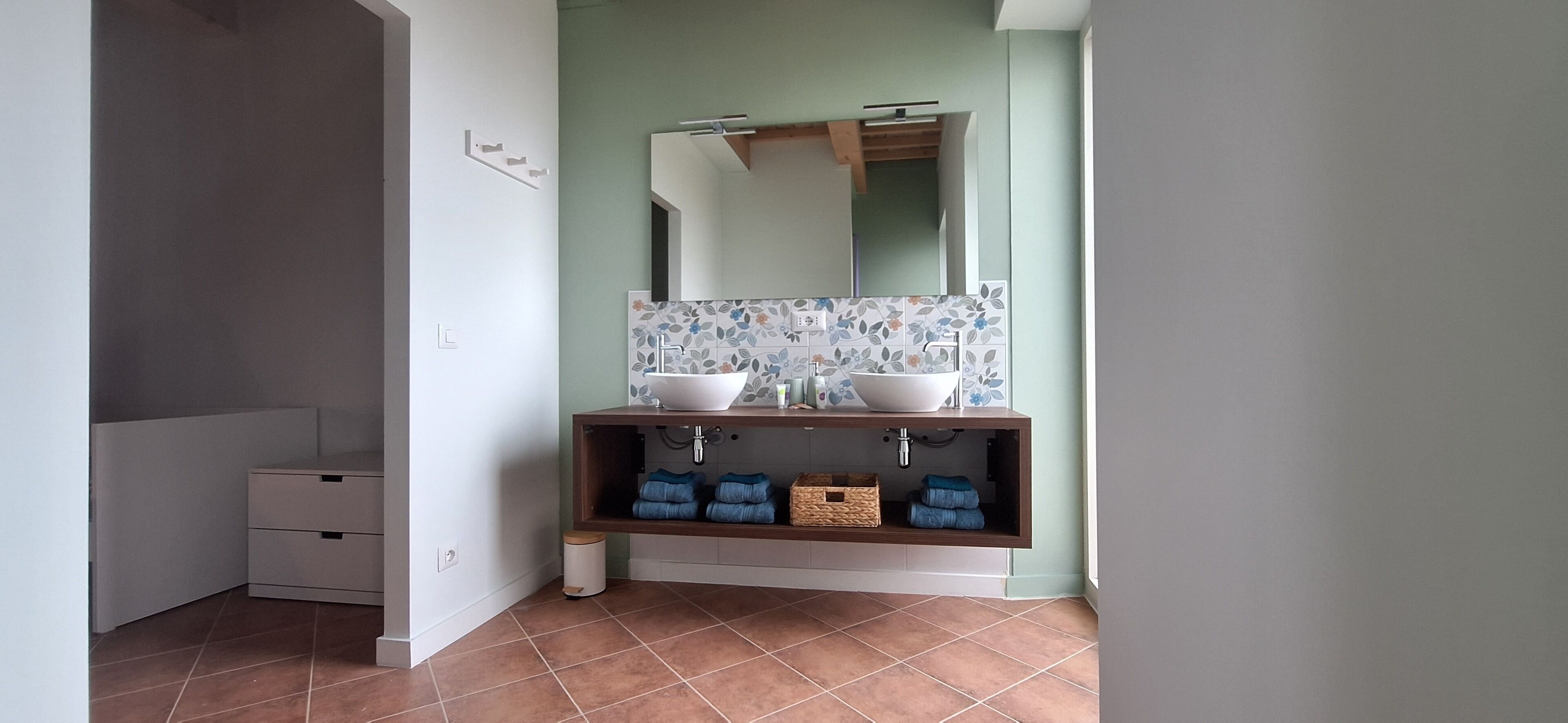 A bathroom with two sinks, a mirror, and a tiled backsplash. There are two towel racks below the sinks and a small trash bin to the left.