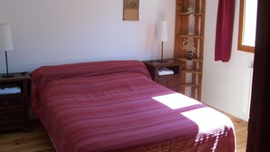 Un dormitorio con cama, mesita de noche, repisa de madera con un florero y ventana con cortinas.