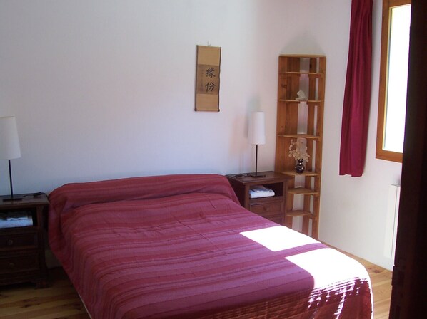 Un dormitorio con cama, mesita de noche, repisa de madera con un florero y ventana con cortinas.