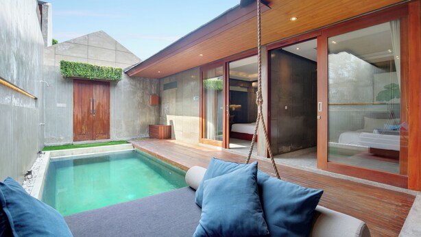 A modern outdoor pool area with a wooden deck, a swing, and a seating area with cushions. - Aqilah Villa (Legian)