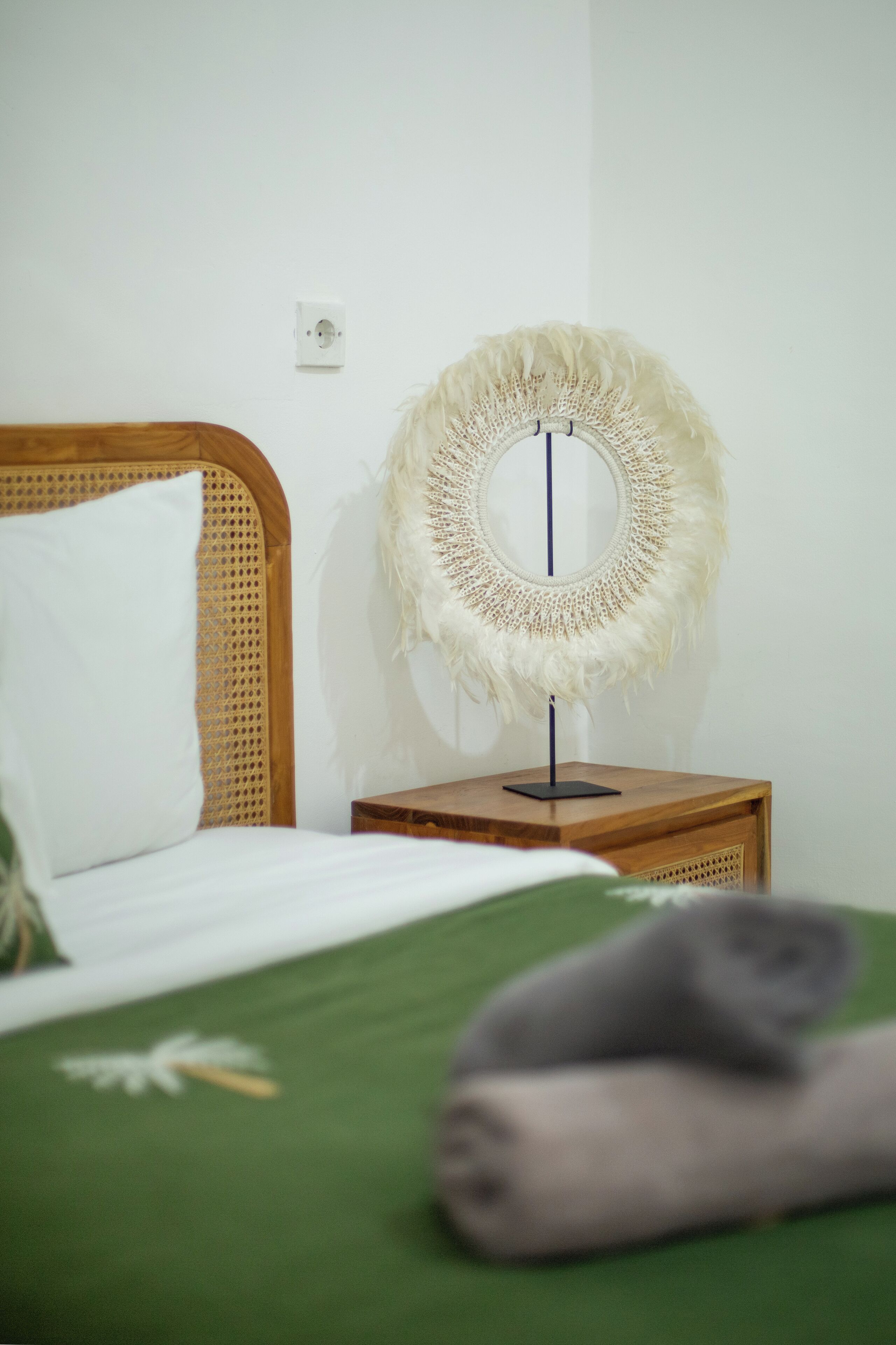 A bed with white bedding, a wooden headboard, a round mirror on a wooden nightstand, and a green area rug.