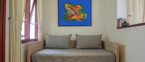 A wicker sofa with grey cushions, a colorful abstract painting on the wall, and a small shelf with books and a flower.