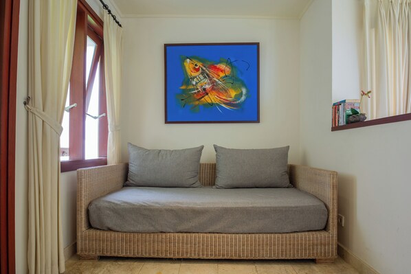 A wicker sofa with grey cushions, a colorful abstract painting on the wall, and a small shelf with books and a flower. - Soleia Surf Camp (Tibubeneng)