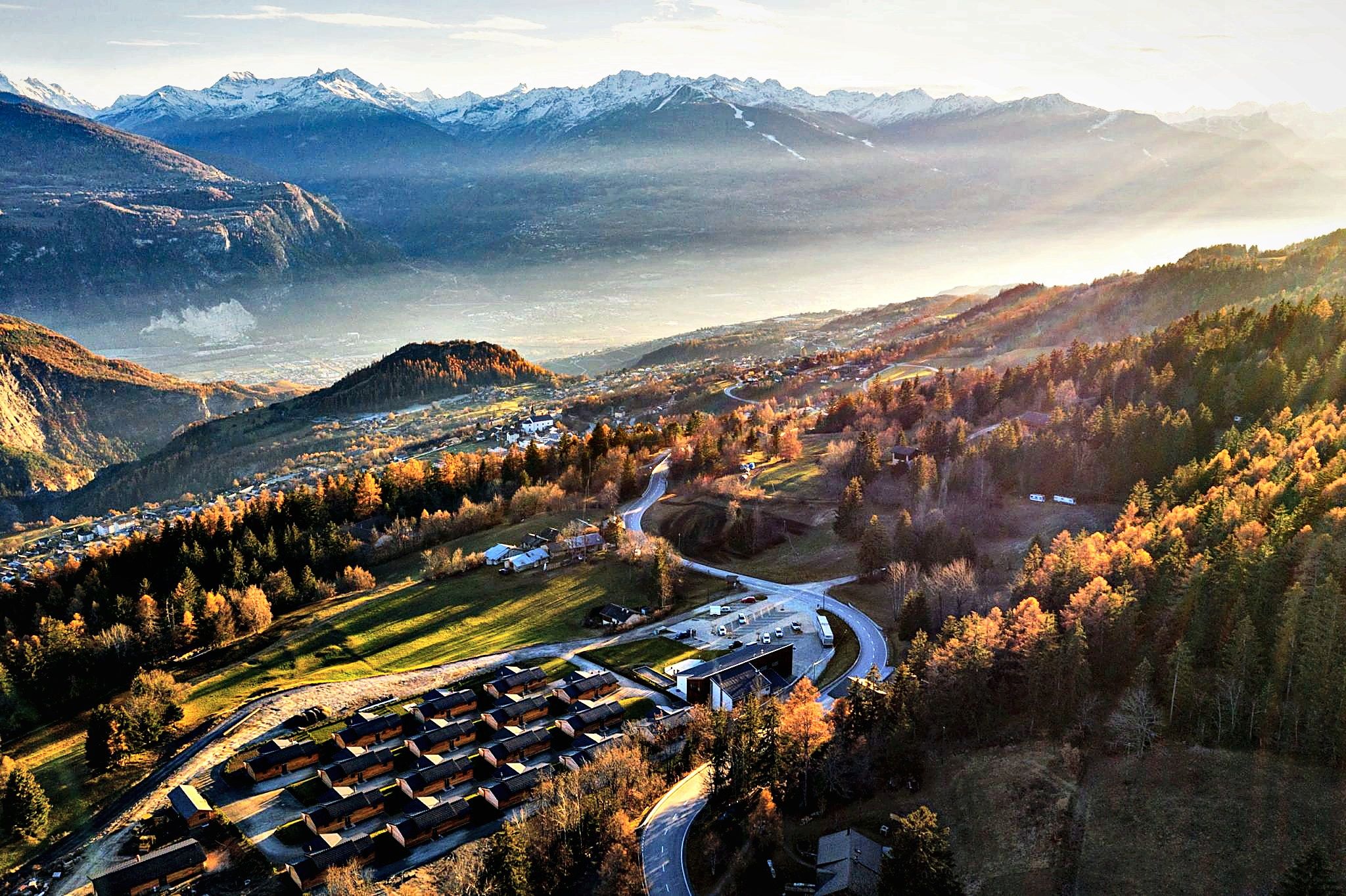 A mountainous landscape with a valley, a winding road, and a cluster of buildings.