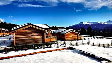 A wooden cabin with a snow-covered roof, surrounded by a snowy landscape and mountains in the background.