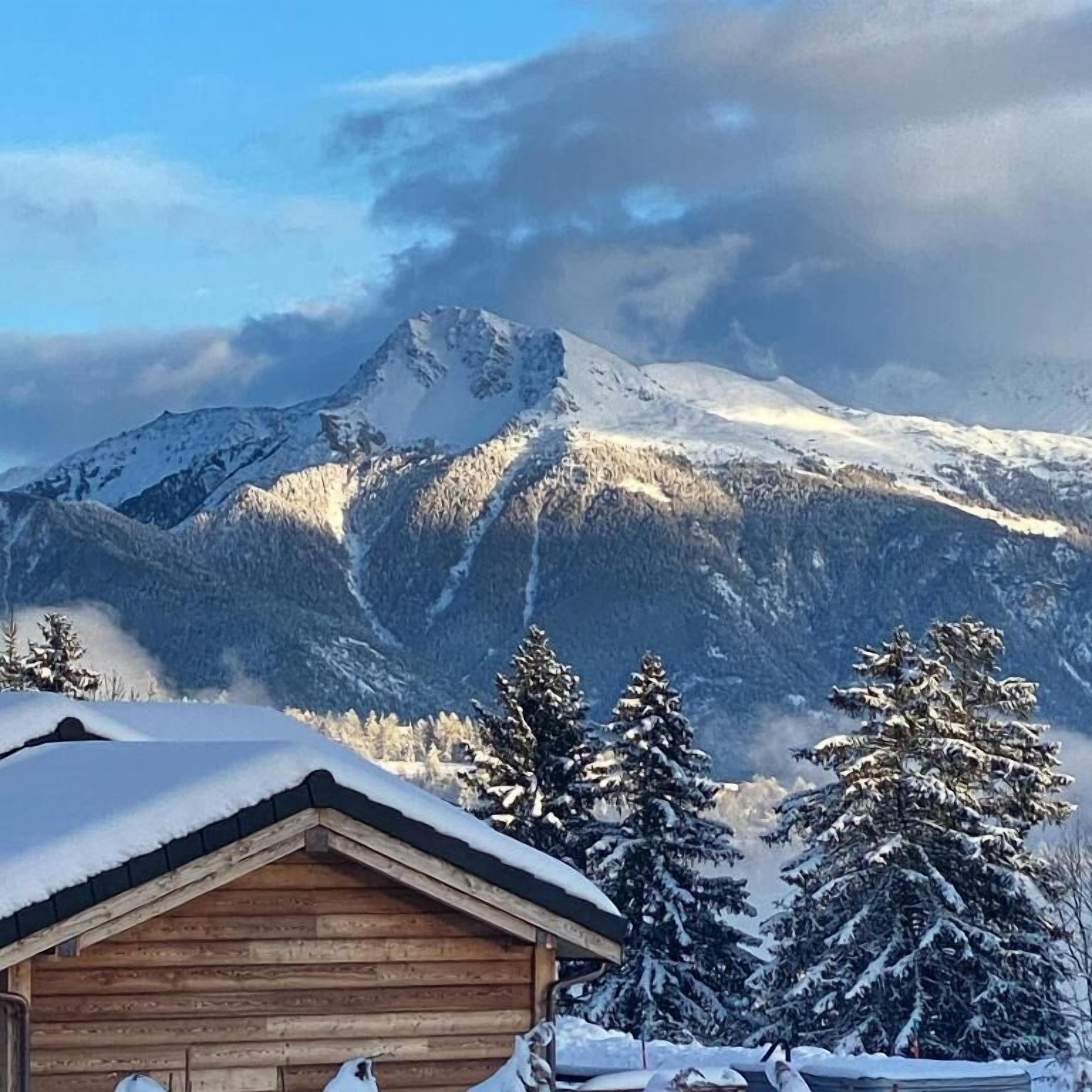 A wooden cabin with a snow-covered roof, surrounded by snow-laden trees and mountains in the background.