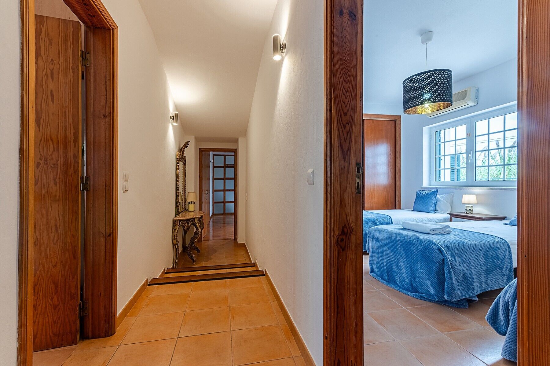 A hallway leading to a bedroom with a bed, a window, and a lamp.