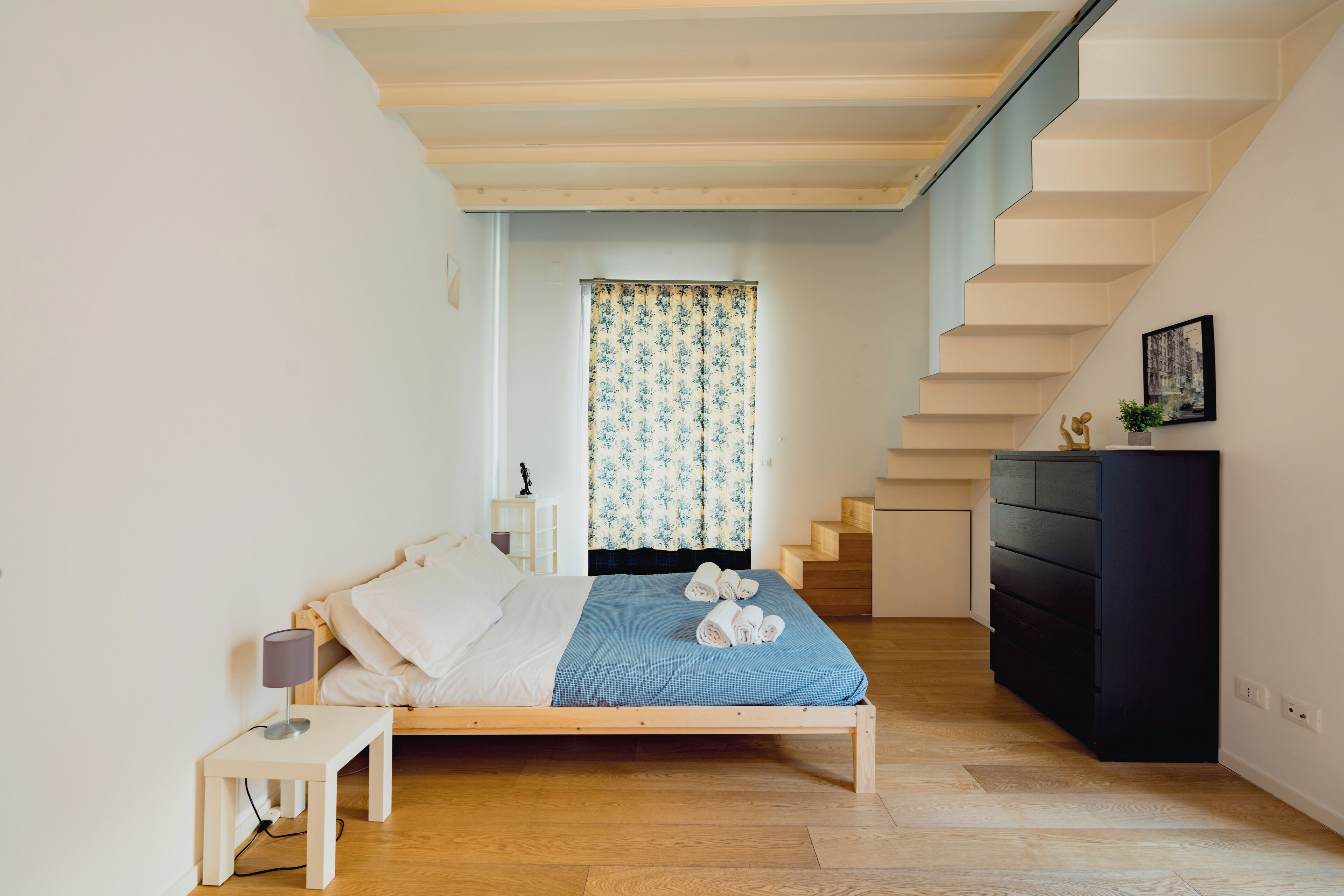 A bedroom with a bed, a bedside table, a dresser, a window with curtains, and a staircase.