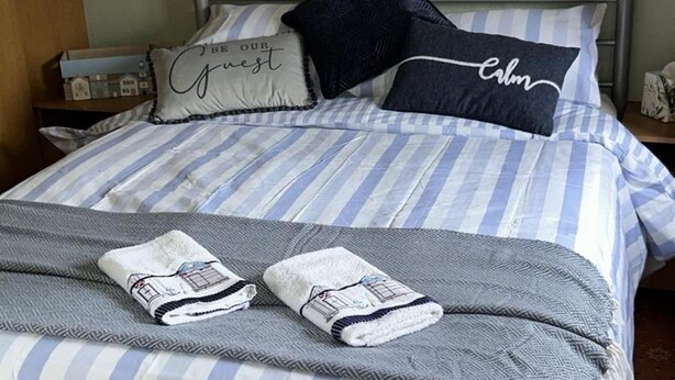 Una cama bien tendida con ropa de cama a rayas y dos toallas, un cuadro en la pared y una mesita de noche de madera con una lámpara.