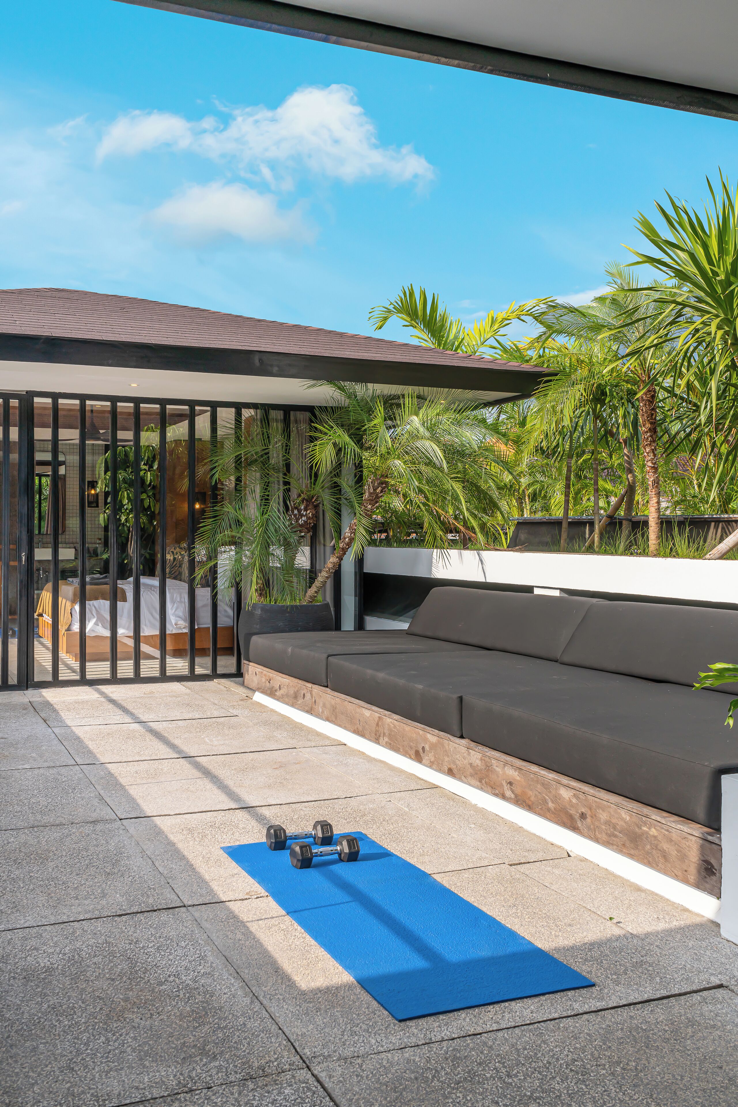 Um espaço ao ar livre moderno com um tapete de exercícios azul, um sofá preto e uma cama visível através de uma divisória de vidro.