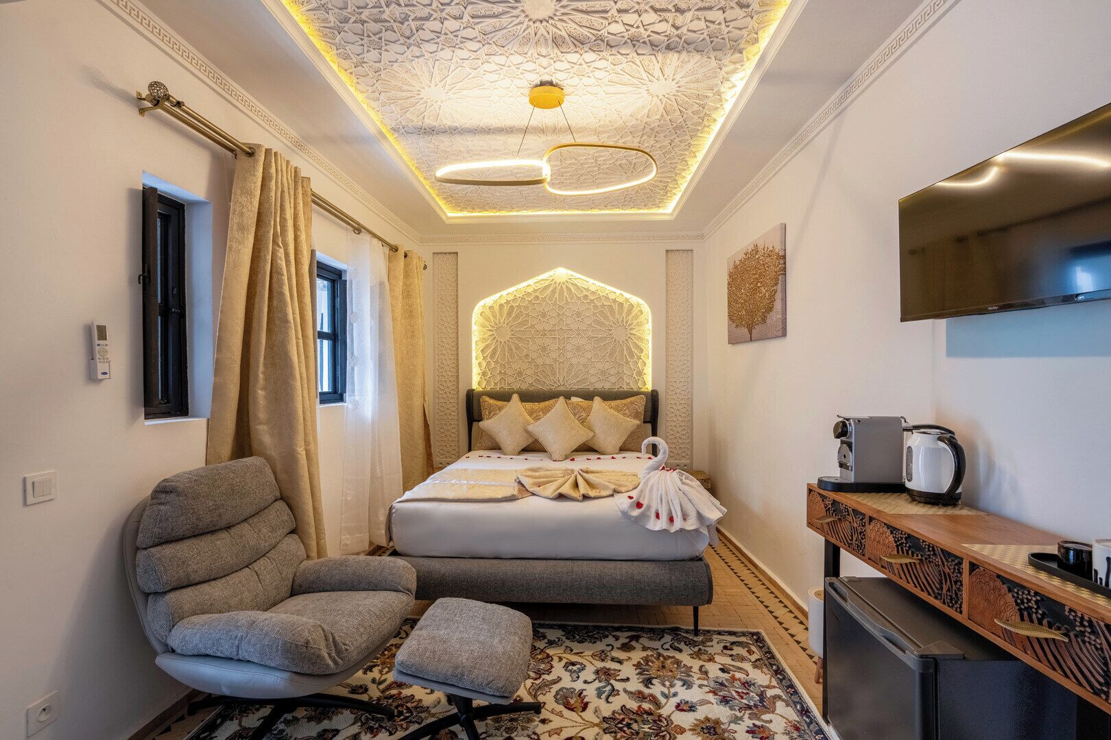 A modern hotel room with a bed, a seating area, a kitchenette, and a decorative ceiling.