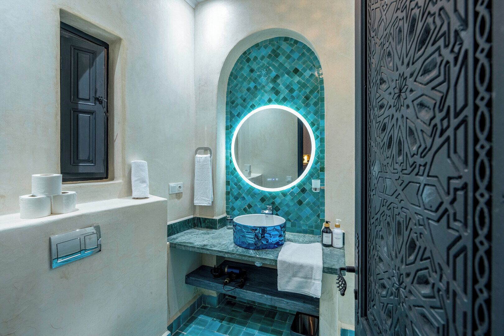 Une salle de bain avec un mur recouvert de mosaïques bleues, un miroir rond, un lavabo avec un récipient bleu et une porte ornée de motifs complexes.