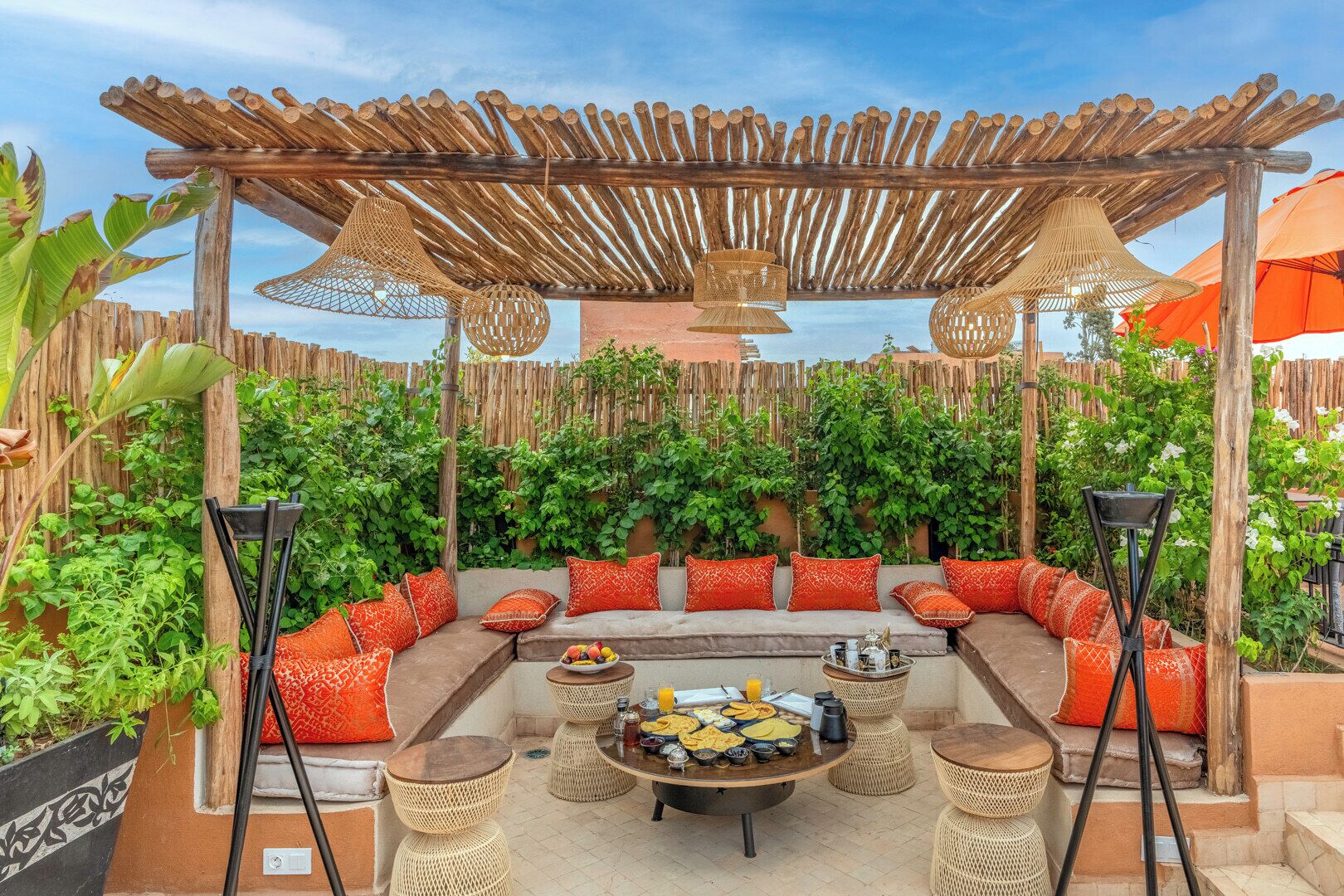 Un espace extérieur abrité, aménagé avec une pergola en bois, des chaises en osier et une table ronde centrale où sont disposés des plats et des boissons.