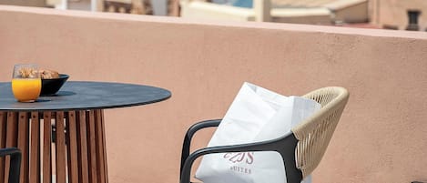 Una mesa redonda con un tazón de cereal y jugo de naranja, una silla de mimbre con cojín blanco, y una vista de edificios y el mar al fondo.