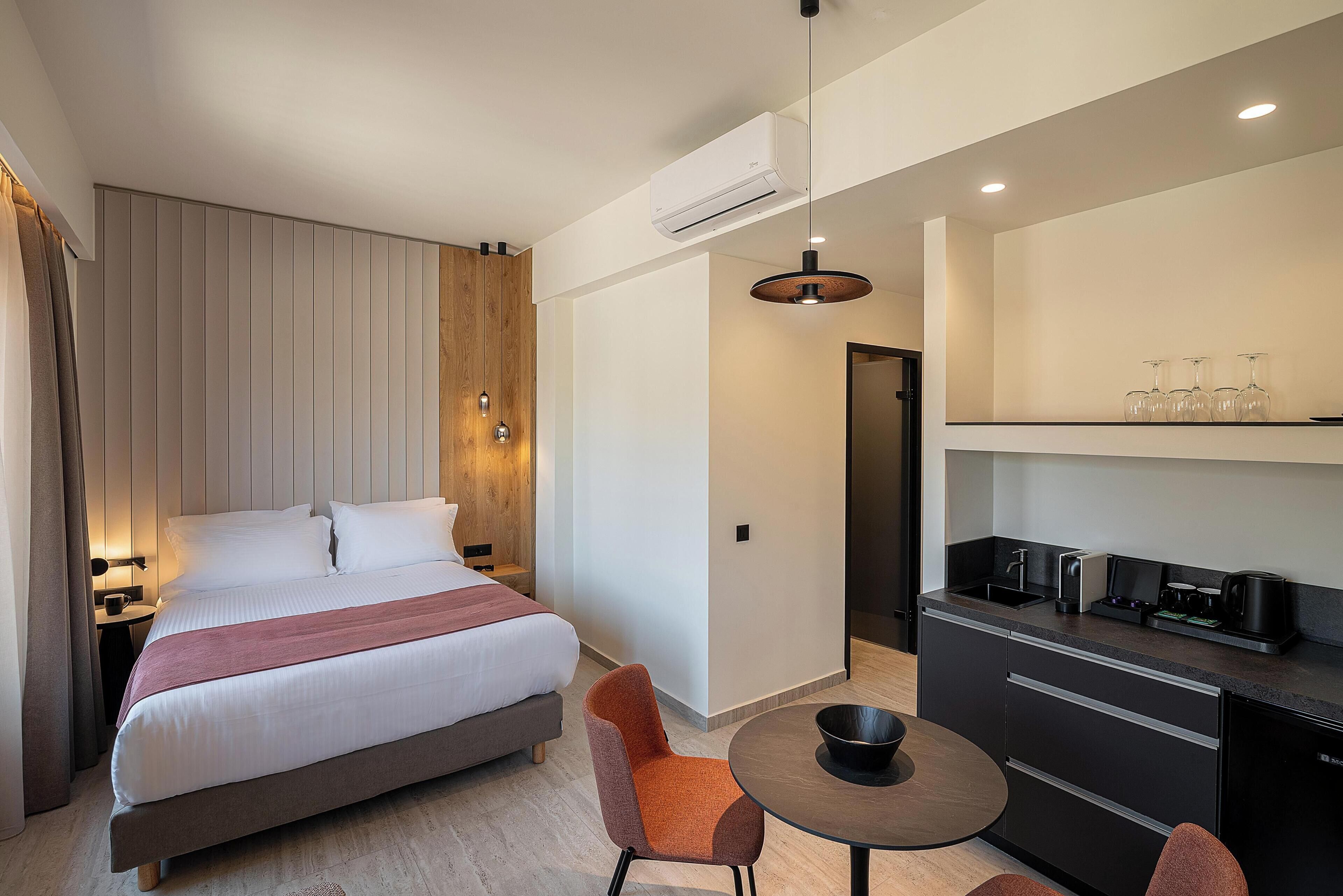 A modern hotel room with a large bed, a dining table, and a kitchenette.
