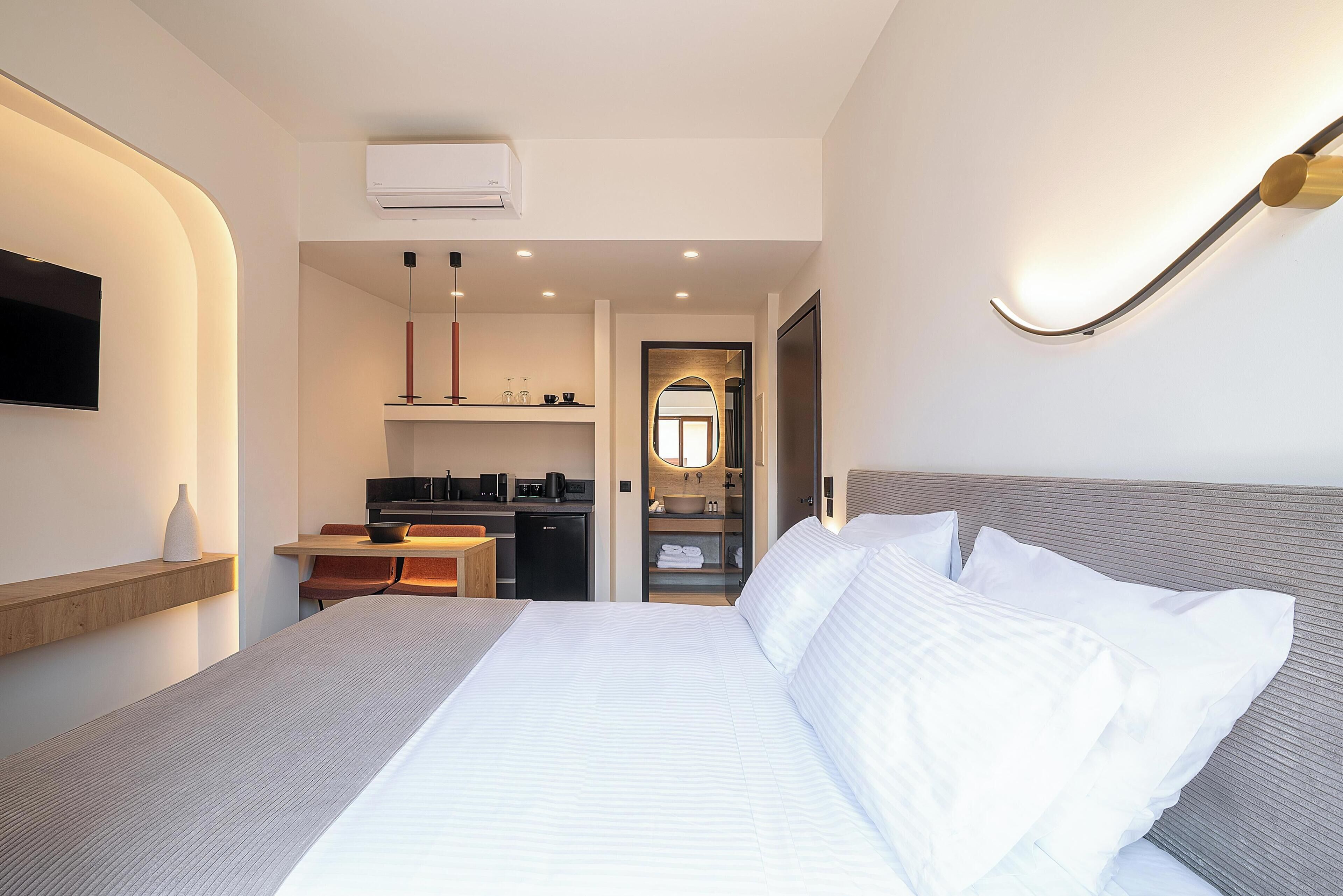 A modern hotel room with a large bed, a desk with a computer, a small kitchenette, and a bathroom visible through an archway.