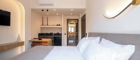 A modern hotel room with a large bed, a desk with a computer, a small kitchenette, and a bathroom visible through an archway.