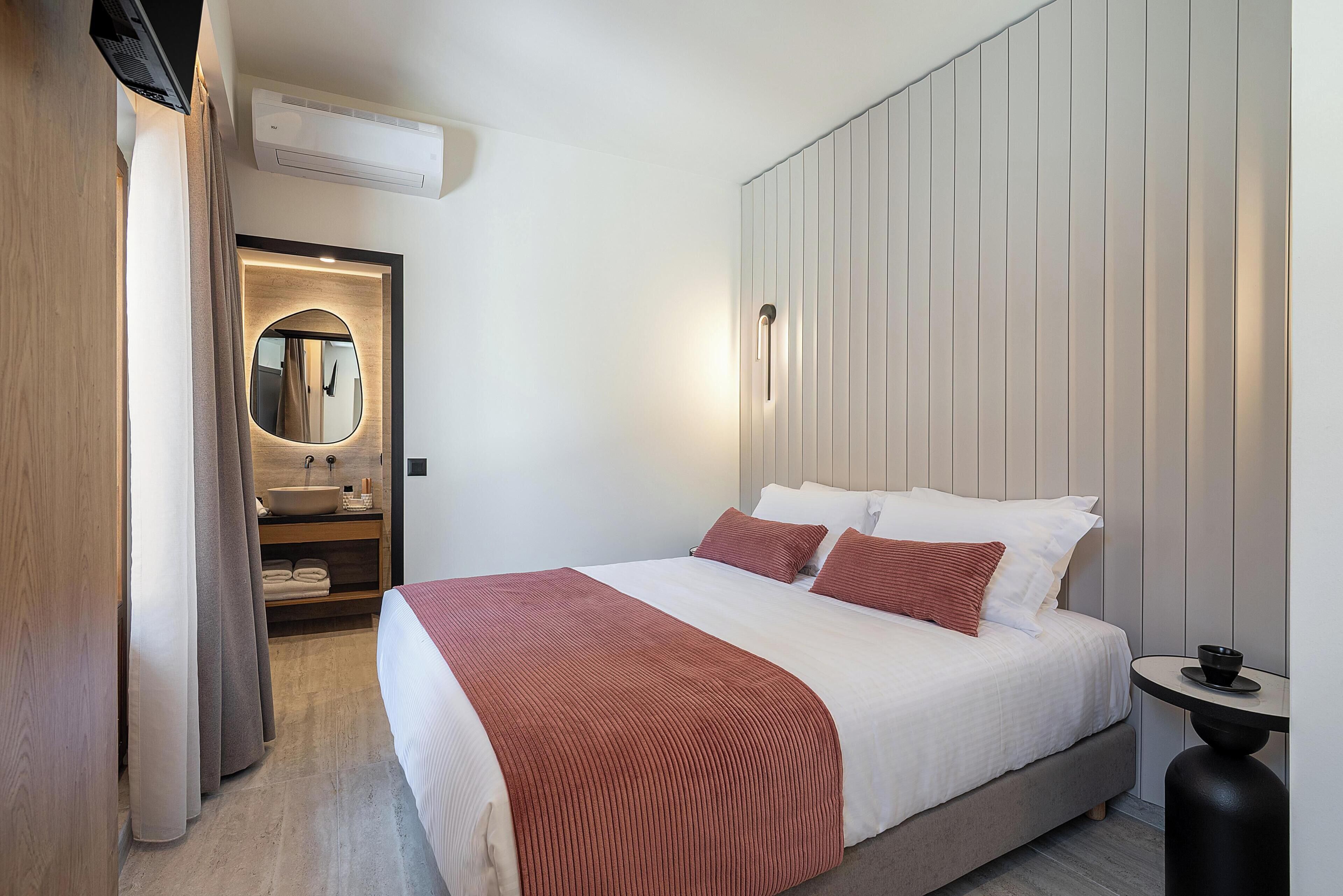 A modern hotel room with a large bed, a bedside table, a built-in shelf with towels, and a mirror.