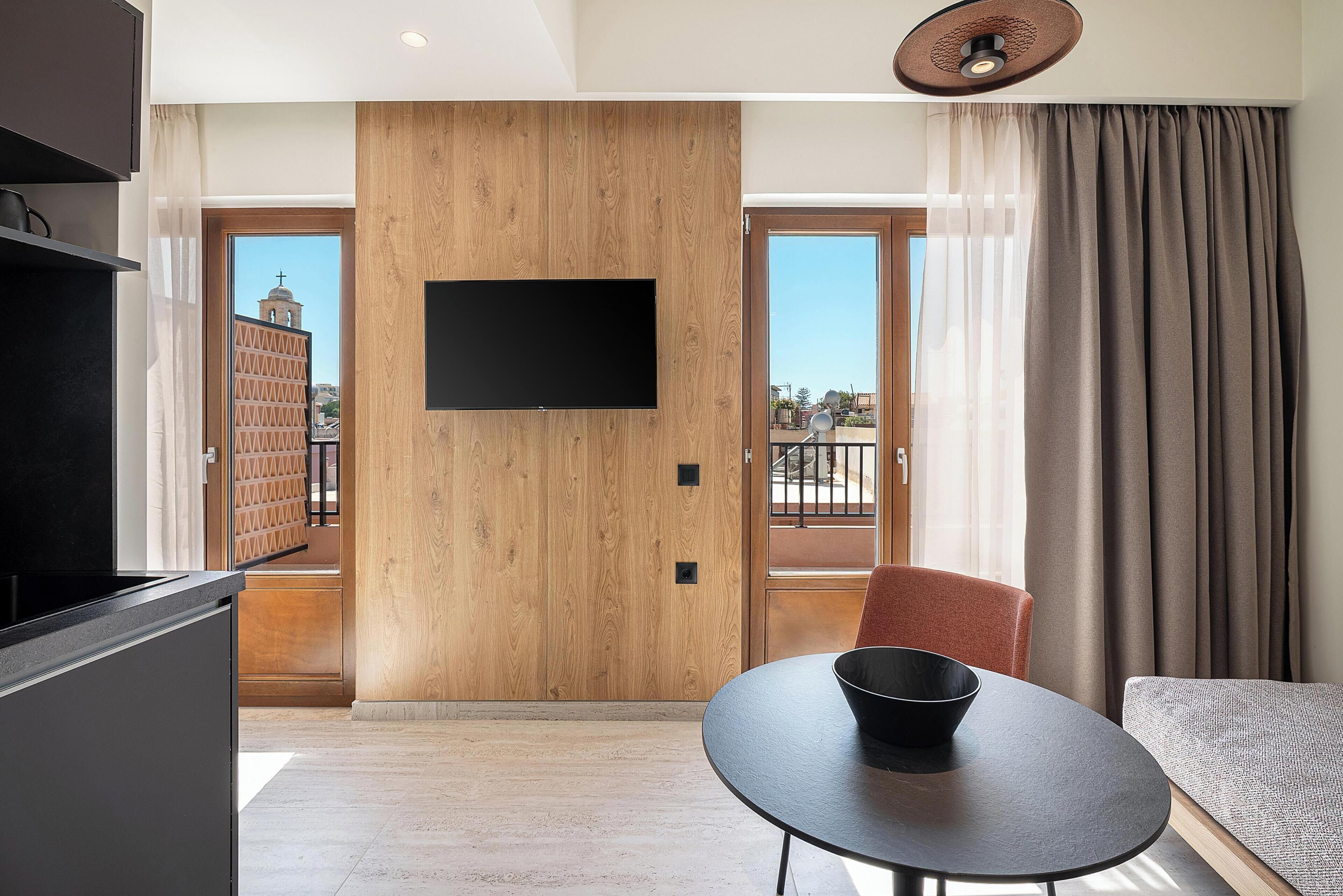 Una habitación de hotel moderna con una pared de acento de madera, un televisor de pantalla plana, una mesa redonda con un cuenco, una silla roja y un amplio ventanal con vistas a un edificio.